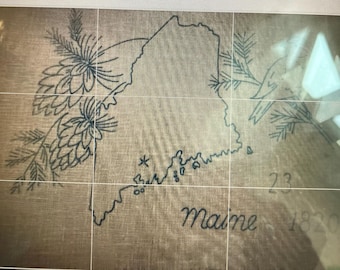 May include: A tan linen fabric featuring a dark blue embroidered outline of the state of Maine. The design includes pine cones and needles on the left, a small bird on the right, and the text "Maine 1820" with the number "23" embroidered below.
