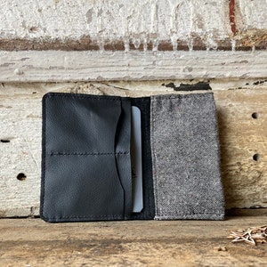 Puede incluir: Una cartera de cuero negro con una solapa de tela gris. La cartera tiene múltiples ranuras para tarjetas y un bolsillo para efectivo.