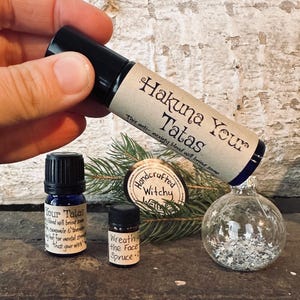 Può includere: Un assortimento di bottiglie di oli essenziali e un ornamento di vetro trasparente pieno di glitter argentati. Una bottiglia recita "Hakuna Your Tatas", un'altra "Your Tatas" e una terza "Wreath the Face". Un piccolo cartello di legno recita "Handcrafted Witchy Ways".