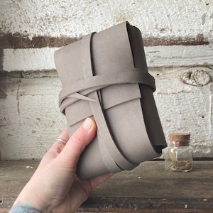 May include: A grey leather journal with a woven strap closure. The journal is wrapped in a grey leather band and tied with a grey leather strap.