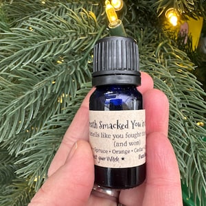 以下が含まれることがあります： 黒いキャップが付いた、小さな濃い青色のエッセンシャルオイルボトル。「Death Smacked You in」と書かれたラベルには、トウヒ、オレンジ、杉などの香りが記載されています。ボトルは緑のクリスマスツリーの前に置かれています。