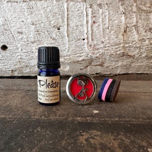 Puede incluir: Una botella de vidrio azul de aceite esencial con tapa negra y una etiqueta que dice "Please". Un medallón de aromaterapia plateado con interior rojo y diseño de gato. Una pila de almohadillas de fieltro de colores.
