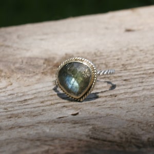 Op de afbeelding: Een zilveren ring met een goudkleurige bezel en een grote, vierkant geslepen, iriserende edelsteen. De edelsteen heeft tinten groen, blauw en grijs. De ringband heeft een gedraaid touwontwerp.