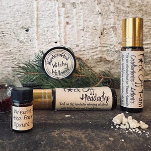 Puede incluir: Una variedad de productos de bienestar artesanales, que incluyen mezclas de aceites esenciales para aliviar el dolor de cabeza y la artritis. La imagen presenta botellas roll-on, un frasco pequeño y una etiqueta circular con el texto "Handcrafted Witchy Wellness."