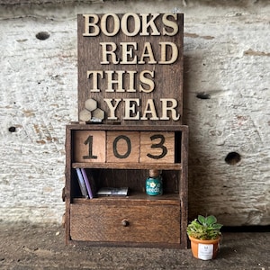 Può includere: Una libreria in legno in miniatura con la scritta "BOOKS READ THIS YEAR" in alto. La libreria mostra il numero "103" su blocchi di legno, un piccolo barattolo con l'etichetta "seeds" e una pianta in vaso in miniatura. Lo schema dei colori è marrone.