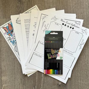 Puede incluir: Una colección de páginas de papel blanco con ilustraciones florales y de mariposas, y texto. Una caja de lápices de colores Faber-Castell Black Edition está en el centro. Los lápices son negros con puntas coloridas. Las páginas son para el diario y la reflexión del tarot.