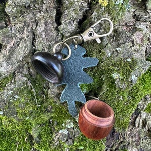 May include: A keyring featuring a dark brown wooden acorn-shaped container with a screw-top lid, attached to a dark green textured leather leaf-shaped charm and a bronze-toned metal lobster clasp hardware for secure attachment to items.