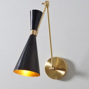 May include: A black and gold wall-mounted light fixture with a cone-shaped shade and a swivel arm. The light is on and illuminating the room.