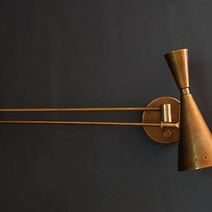 Puede incluir: Una l&aacute;mpara de pared de brazo oscilante de lat&oacute;n con una pantalla c&oacute;nica. La l&aacute;mpara tiene un brazo doble y una base redonda.