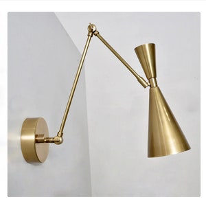 May include: A gold-toned wall sconce with a long, adjustable arm and a cone-shaped shade. The sconce is mounted on a white wall.