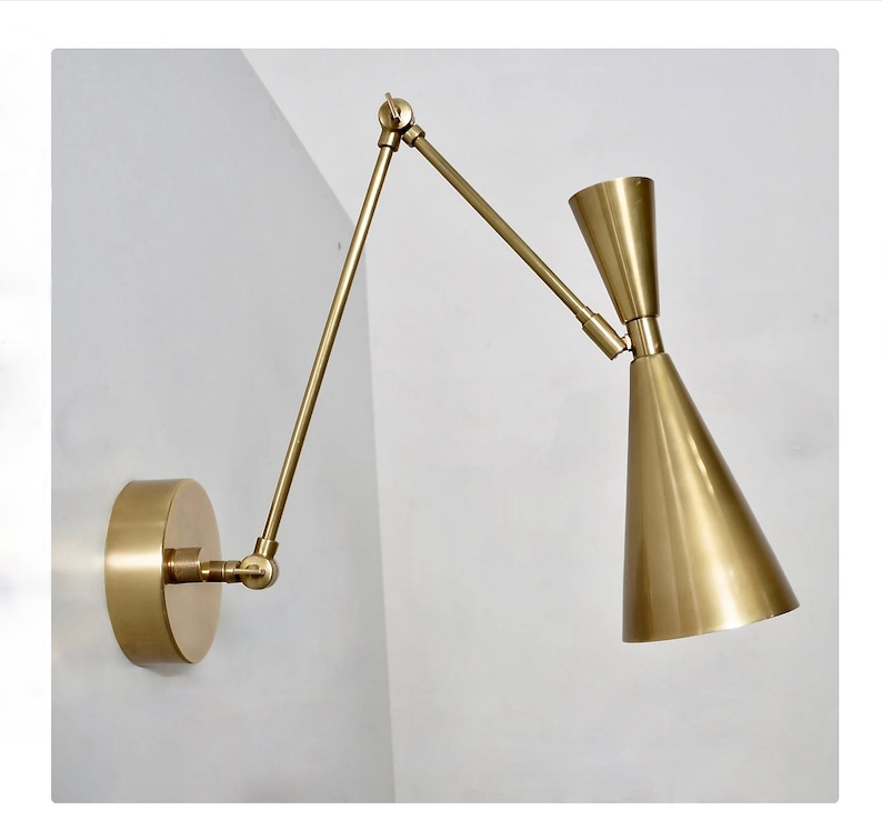 May include: A gold-toned wall sconce with a long, adjustable arm and a cone-shaped shade. The sconce is mounted on a white wall.
