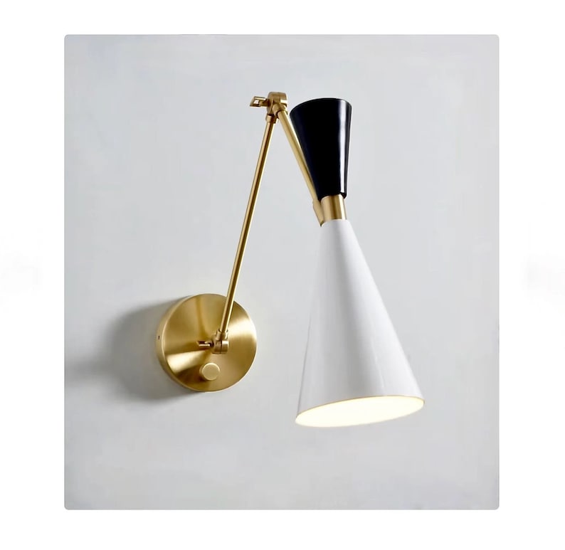 May include: A gold wall-mounted light fixture with a white and black cone-shaped shade. The shade is adjustable and can be angled up or down.