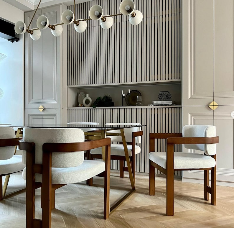 May include: A dining room with a glass table and six chairs. The chairs are upholstered in white fabric and have wooden frames. The table has a brass base. The room has a modern design with a wood slat wall and a light fixture with white globes.