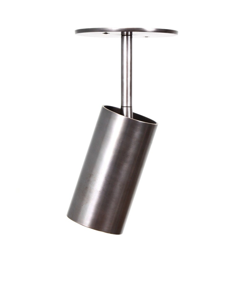 May include: A silver metal ceiling light fixture with a cylindrical shade that is angled downwards. The light fixture has a round base with a small hole in the center.
