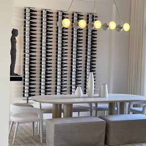 May include: A modern dining room with a long wooden table and white chairs. The wall behind the table features a wine rack with many bottles of wine. A gold chandelier with round light bulbs hangs above the table.