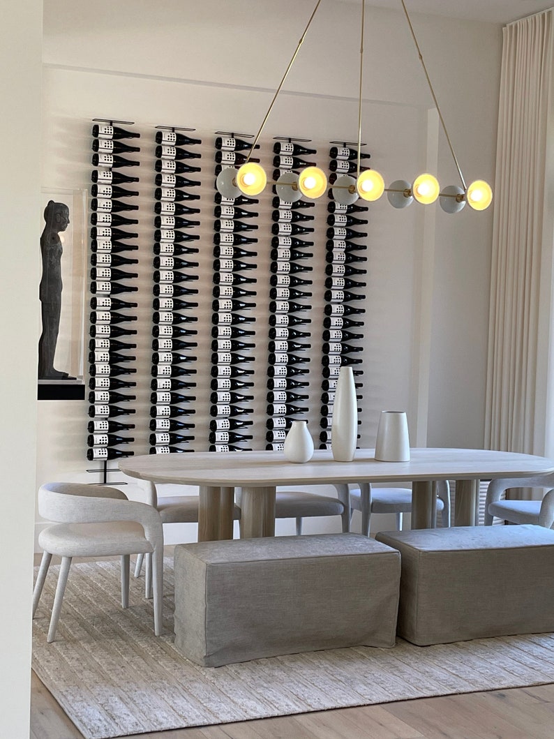 May include: A modern dining room with a long wooden table and white chairs. The wall behind the table features a wine rack with many bottles of wine. A gold chandelier with round light bulbs hangs above the table.