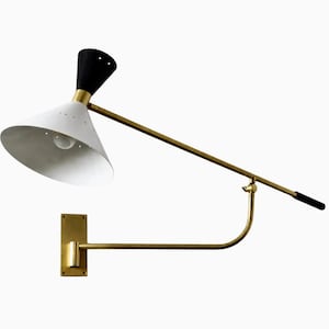May include: A wall-mounted lamp featuring a long, adjustable brass arm with a black handle. The lamp head is a cone-shaped shade with a white interior and exterior, accented by a black top section and a series of small perforations near the rim.
