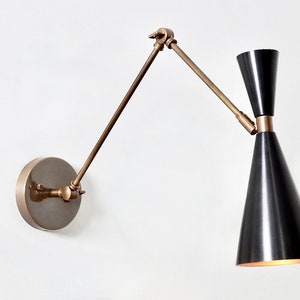 May include: A brass wall-mounted light fixture with a black cone-shaped shade. The arm of the light fixture is adjustable and can be moved up and down.