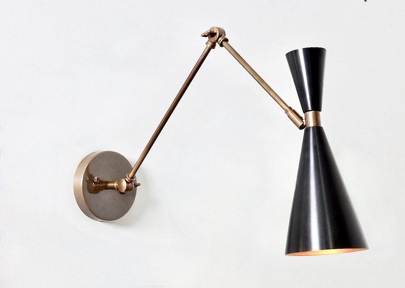 May include: A brass wall-mounted light fixture with a black cone-shaped shade. The arm of the light fixture is adjustable and can be moved up and down.