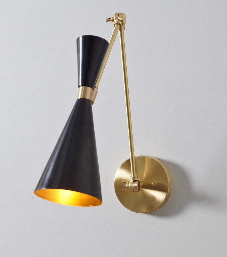 May include: A black and gold wall-mounted light fixture with a cone-shaped shade and a swivel arm. The light is on and illuminating the room.