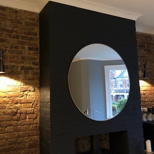 Puede incluir: Un gran espejo redondo cuelga sobre una chimenea negra en una habitaci&oacute;n con paredes de ladrillo visto. Dos apliques de pared negros y dorados con una pantalla en forma de cono iluminan la habitaci&oacute;n.