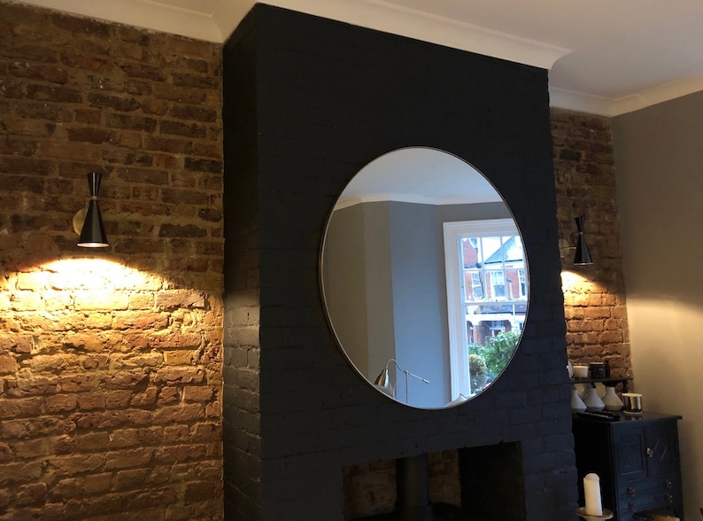 Puede incluir: Un gran espejo redondo cuelga sobre una chimenea negra en una habitaci&oacute;n con paredes de ladrillo visto. Dos apliques de pared negros y dorados con una pantalla en forma de cono iluminan la habitaci&oacute;n.