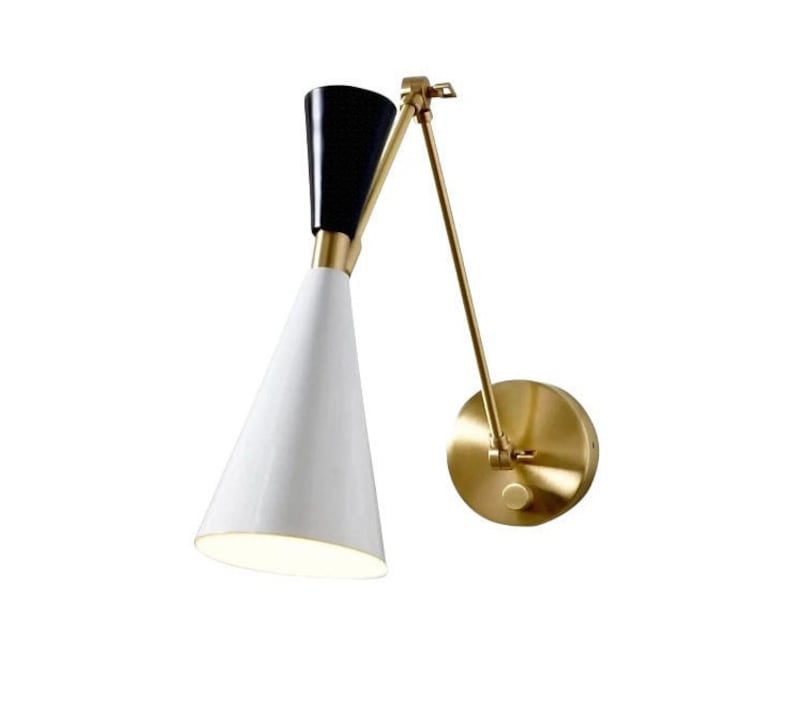 May include: A gold-toned wall sconce with a white and black cone-shaped shade. The sconce has a flexible arm that can be adjusted to direct the light.
