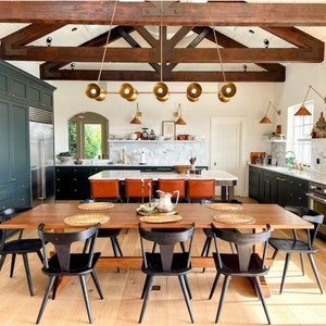 May include: A rustic kitchen with dark green cabinets, a large wooden table with black chairs, and a chandelier with gold light fixtures. The kitchen features exposed wooden beams and a marble countertop.