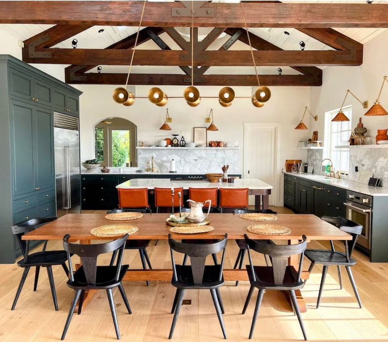 May include: A rustic kitchen with dark green cabinets, a large wooden table with black chairs, and a chandelier with gold light fixtures. The kitchen features exposed wooden beams and a marble countertop.