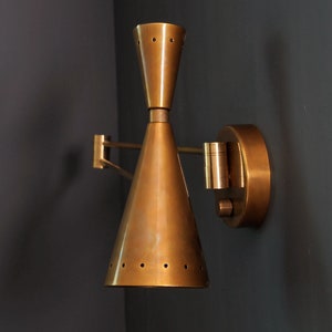 Puede incluir: Una l&aacute;mpara de pared de lat&oacute;n con una pantalla c&oacute;nica y un brazo ajustable. La pantalla tiene una serie de peque&ntilde;os agujeros alrededor del borde inferior.