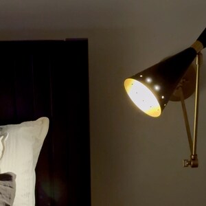 May include: A black and gold wall-mounted lamp with a cone-shaped shade. The lamp is on and casting a warm light.