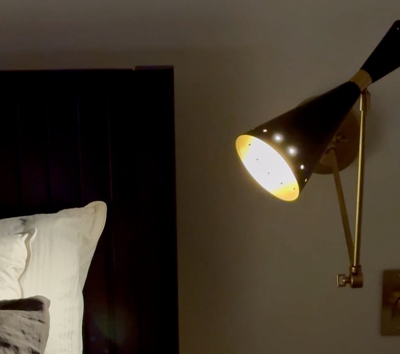 May include: A black and gold wall-mounted lamp with a cone-shaped shade. The lamp is on and casting a warm light.