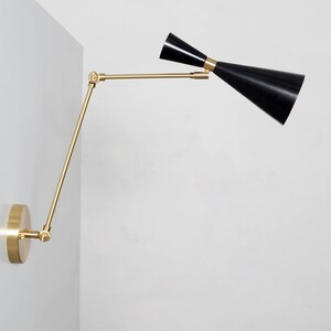 May include: A brass wall-mounted light fixture with a black adjustable shade. The shade is angled upwards and the arm is extended outwards.