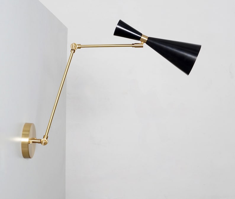 May include: A brass wall-mounted light fixture with a black adjustable shade. The shade is angled upwards and the arm is extended outwards.