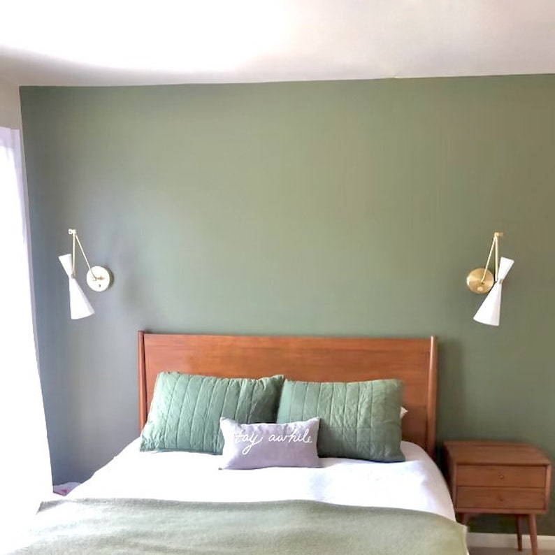 May include: A bedroom with a green wall, a wooden bed frame, and a bedside table. The bed is made with white sheets and a green and white patterned throw blanket. There is a pillow with the text "stay awhile" on it. The room has two gold wall sconces with white shades.