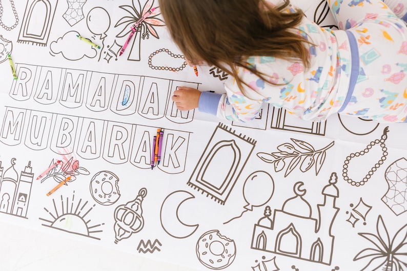May include: A white coloring sheet with the words "Ramadan Mubarak" in a banner design. The sheet is filled with various Islamic symbols, including a mosque, crescent moon, palm tree, and prayer beads. A young person is coloring the sheet with crayons.