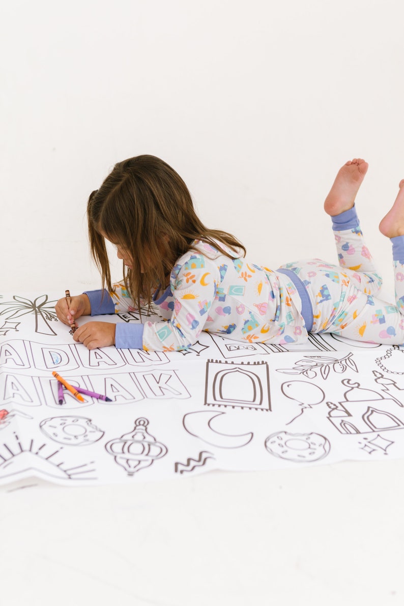 May include: A child in pajamas is coloring a large sheet of paper with black and white illustrations of Islamic symbols and the words "Ramadan Mubarak".