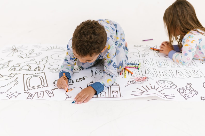 May include: Two children in pajamas color a large white sheet with black outlines of Islamic symbols and the words "Eid Mubarak".
