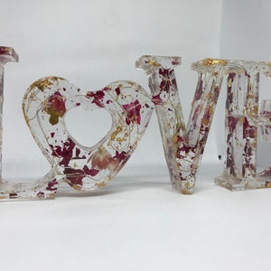 K&ouml;nnte beinhalten: Transparente Harzbuchstaben, die "LOVE" buchstabieren, mit einem herzf&ouml;rmigen "O". Die Buchstaben sind mit getrockneten Blumen und Blattgold gef&uuml;llt.