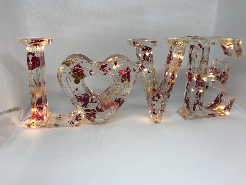 K&ouml;nnte beinhalten: Ein klares Harz-Schild "LOVE" mit eingebetteten getrockneten Blumen und Blattgold. Die Buchstaben sind mit kleinen wei&szlig;en Lichtern beleuchtet.