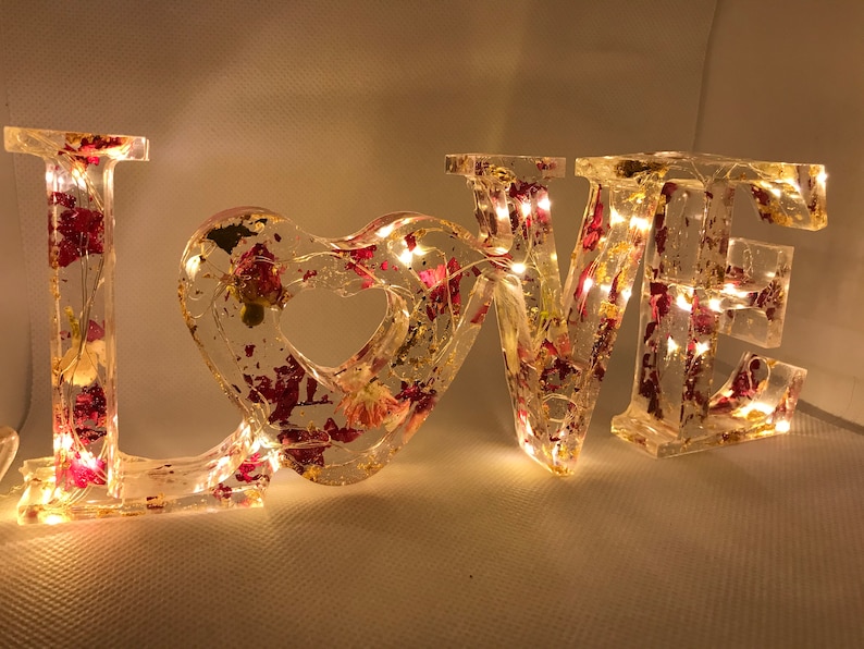 K&ouml;nnte beinhalten: Ein klares Harz-Schild "LOVE" mit eingebetteten getrockneten Blumen und Blattgold. Das Schild wird mit warmwei&szlig;en LED-Lichtern beleuchtet.