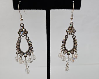 May include: A pair of silver dangle earrings with clear iridescent crystals. The earrings feature a teardrop-shaped design with a small open circle in the center.
