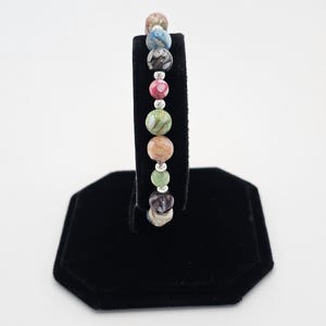 May include: A beaded bracelet featuring a variety of colorful, round gemstones. The bracelet includes beads in shades of pink, green, blue, and brown, separated by small silver-colored beads. The bracelet is displayed on a black stand against a white background.
