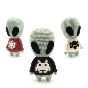 May include: Three crocheted alien toys wearing different sweaters. The first alien is wearing a red sweater with a white snowflake design. The second alien is wearing a black sweater with a white pixelated alien design. The third alien is wearing a white sweater with a black skull and crossbones design.