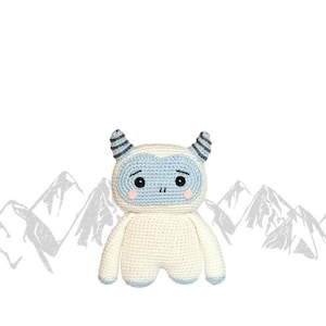 May include: A white crocheted yeti with blue eyes and a light blue face. The yeti has small, grey horns and is standing in front of a grey mountain range.