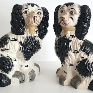 May include: Two ceramic dog ornaments. The dogs are seated, with black and white speckled bodies, black floppy ears, and tan collars. The dogs have detailed facial features, including brown eyes and painted noses. The ornaments are decorative.
