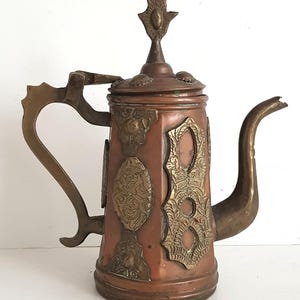 May include: An antique copper and brass teapot with intricate detailing. The teapot has a hinged lid, a curved spout, and a large handle. The body is reddish-brown, with brass accents and decorative patterns. A vintage tea pot.