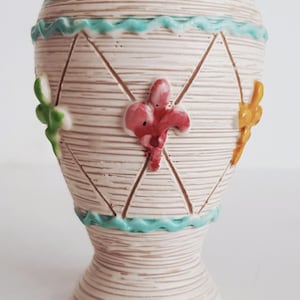 May include: A vintage ceramic vase with a cream-coloured, textured body. It features a turquoise rim and base, with a wavy turquoise band. The vase is adorned with red, green, and yellow floral accents and a diamond pattern.