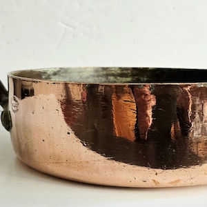 May include: A copper-coloured, oval-shaped pan with a long, dark handle. The pan has a reflective surface, showing the surrounding environment. The handle is attached with a rivet. The pan is likely used for cooking.