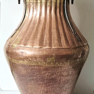 May include: An antique copper vase with a hammered texture. The vase has a wide, rounded body that narrows to a neck and flared rim. It features two handles and a decorative band. The vase is approximately 61 cm tall.